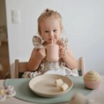 Une petite fille assise à table en train de boire dans un gobelet rose avec une paille